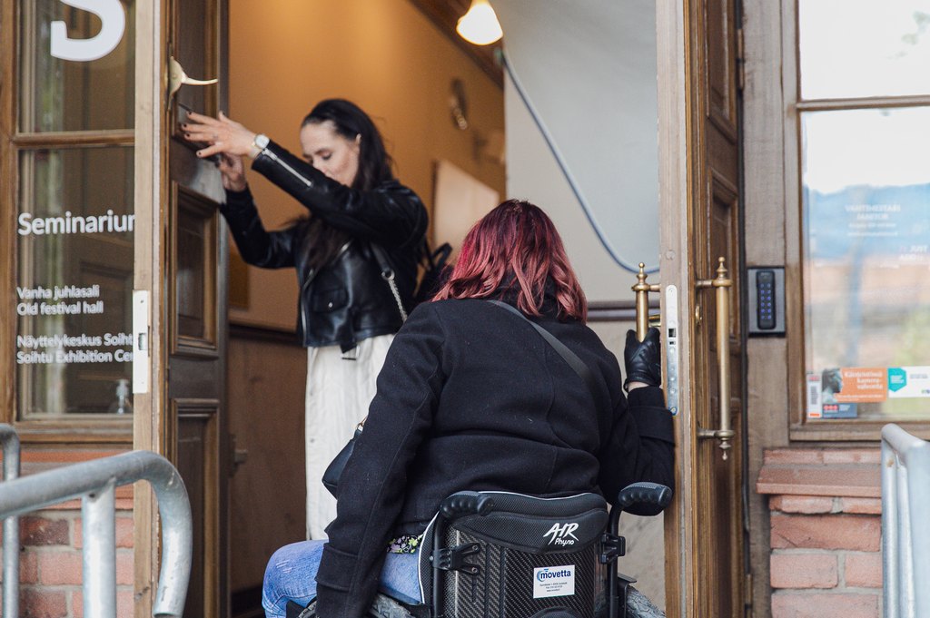 Nainen auttaa pyörätuolilla liikkuvaa opiskelijaa pääsemään liian kapeasta ovesta sisälle