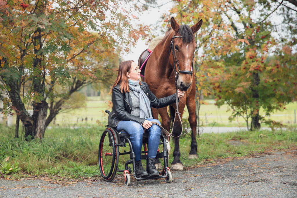 Pyörätuolissa istuva silmälasipäinen nainen pitelee ruskeaa hevosta. Taustalla näkyy syksyisiä puita ja vehreä pelto.
